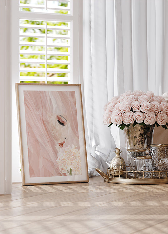 a framed artwork of a woman's face with a pink background, placed on a wooden surface next to a vase of pink roses and a gold tray with a pineapple.